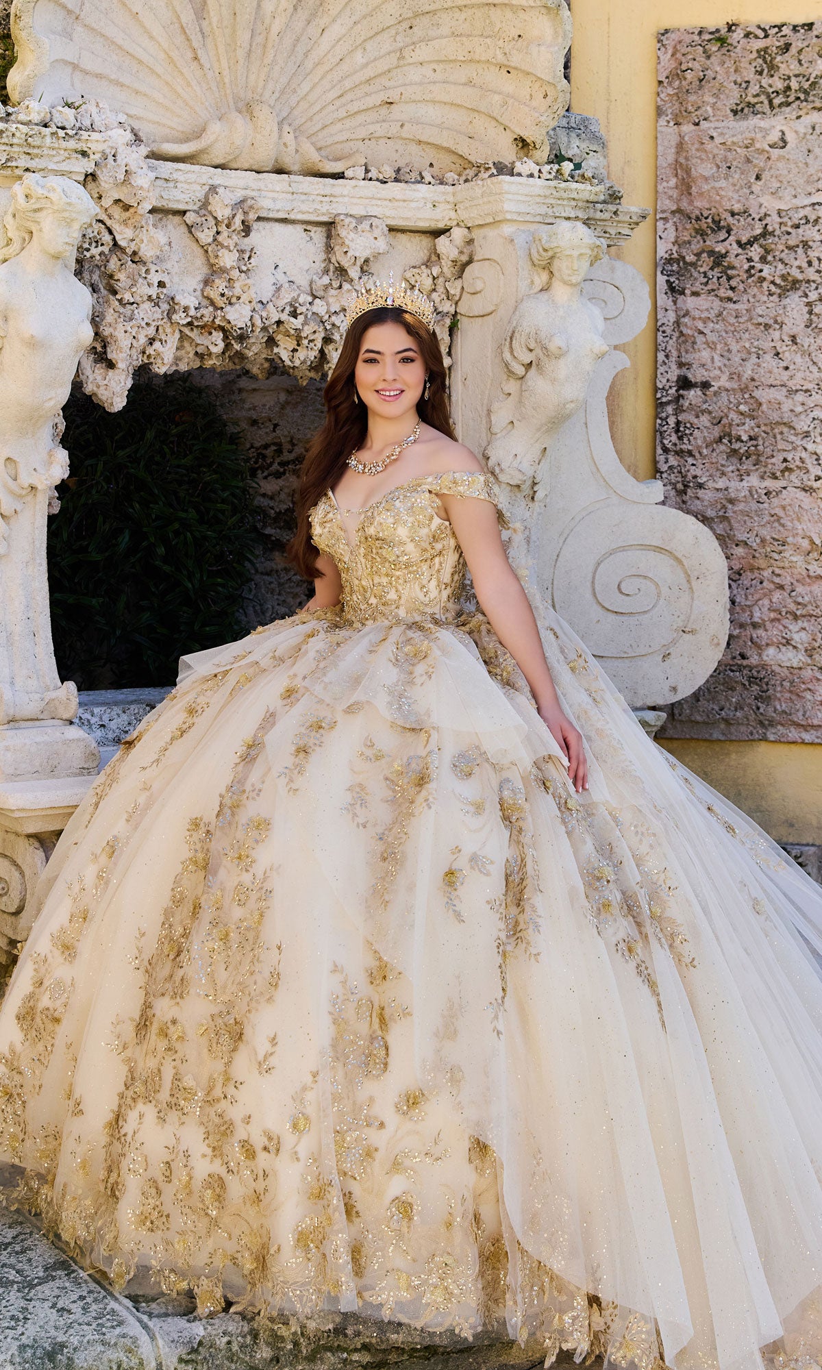 A young woman poses in front of a decorative stone structure wearing the Quinceanera Dress PR30119 By Princesa, featuring an ornate off-the-shoulder design and a detachable cathedral cape.